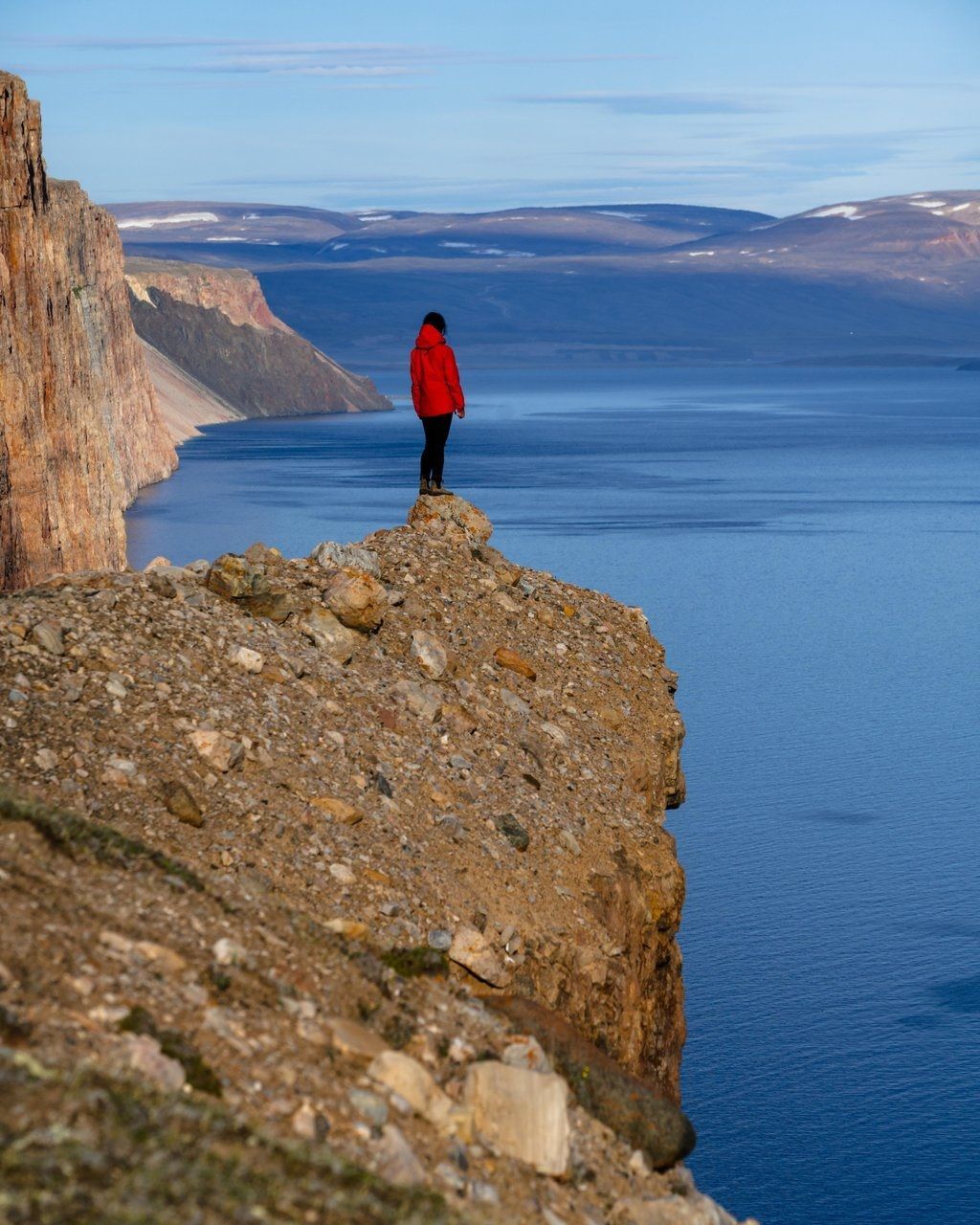 @Arctic Bay Adventures De persoon in de rode jas staat op de rand van een klif en kijkt uit over een uitgestrekt blauw meer en de bergen in de verte.