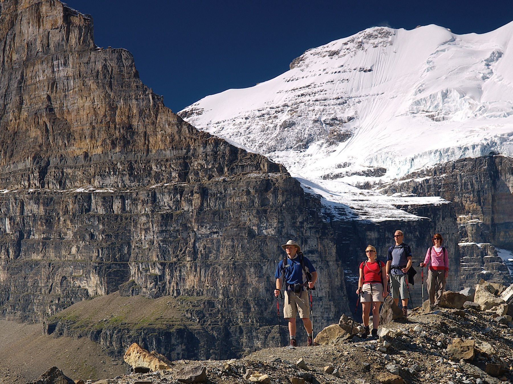 Hikers on a rocky mountain trail with snow-capped peaks in the background under a blue sky.