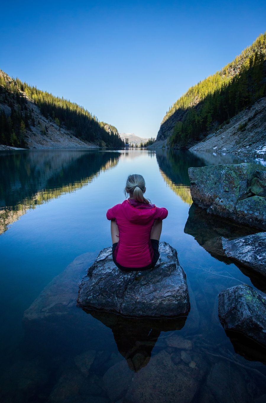 Paul Zizka @paulzizkaphoto / Banff & Lake Louise Tourism (BLLT) Vrouw in rode jas zit op een rots aan het meer, omringd door bergen onder een blauwe hemel.
