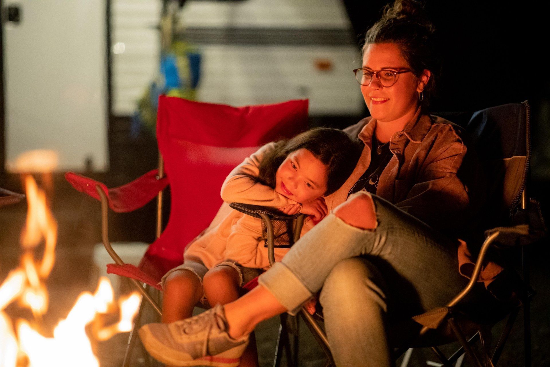 @May Contain Studio-Courtesy of Travel Manitoba Een vrouw en een kind zitten op stoelen bij een kampvuur. Het kind legt zijn hoofd op de schoot van de vrouw en glimlacht. Donkere buitenomgeving.