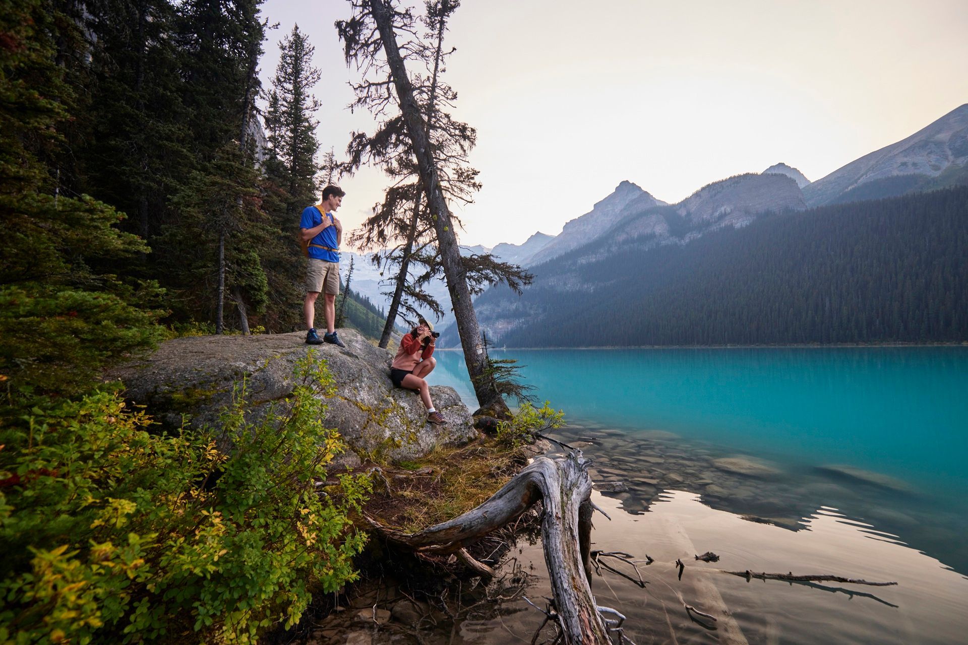 Two people near a turquoise lake, mountains in the background. Man standing, looking at phone. Woman sitting, taking photos.