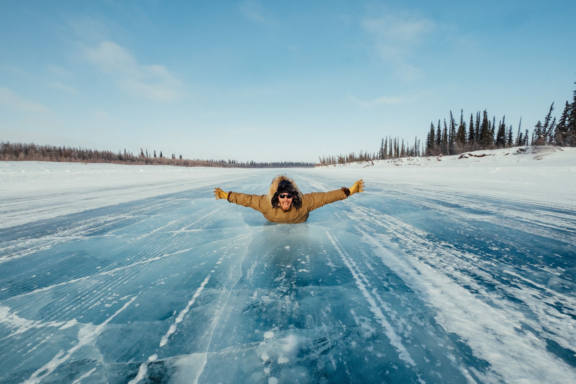 Person with arms outstretched lying on a frozen blue lake. Snowy landscape with trees and bright sky.