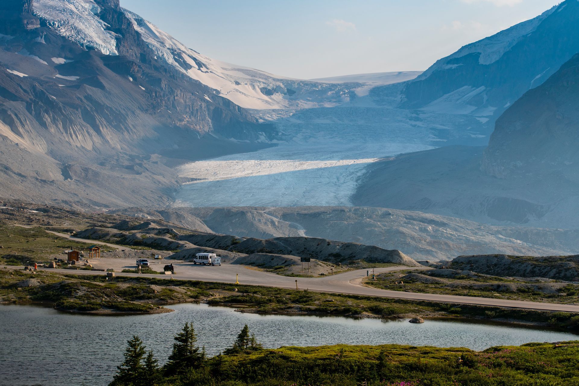 A glacier nestled between mountains, viewed from a lake and a parking area with vehicles.