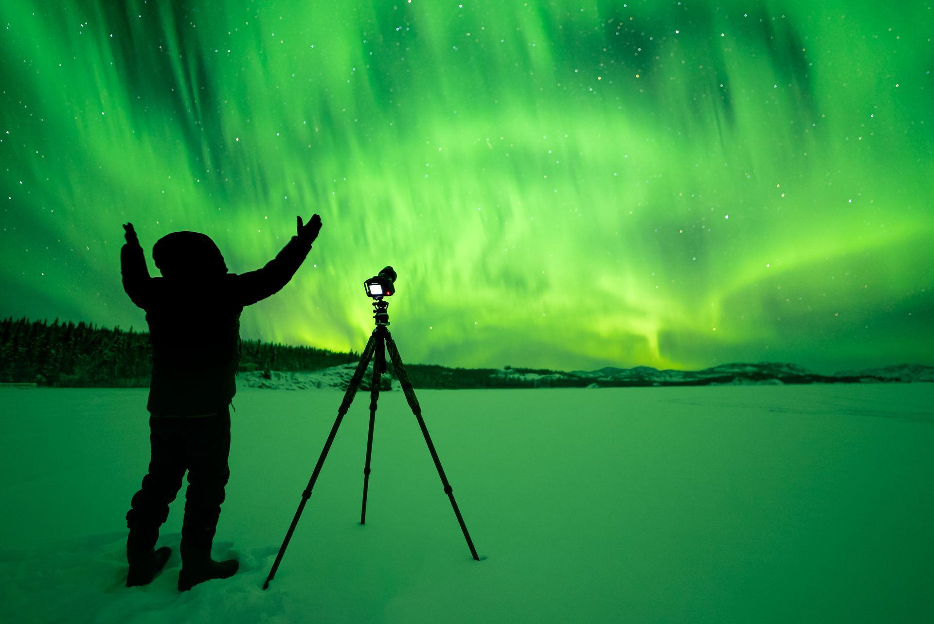 Person silhouetted with arms raised toward vibrant green aurora borealis. Camera on tripod in snowy landscape.