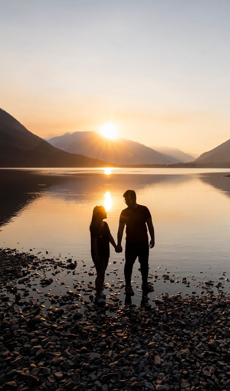 Couple holding hands, silhouetted at the water's edge, watching the sunset over mountains and a lake.