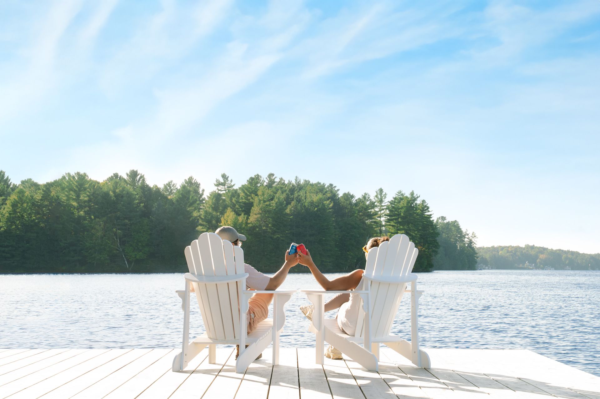Two people clink glasses, relaxing in white chairs on a wooden dock over a lake, with trees and blue sky.