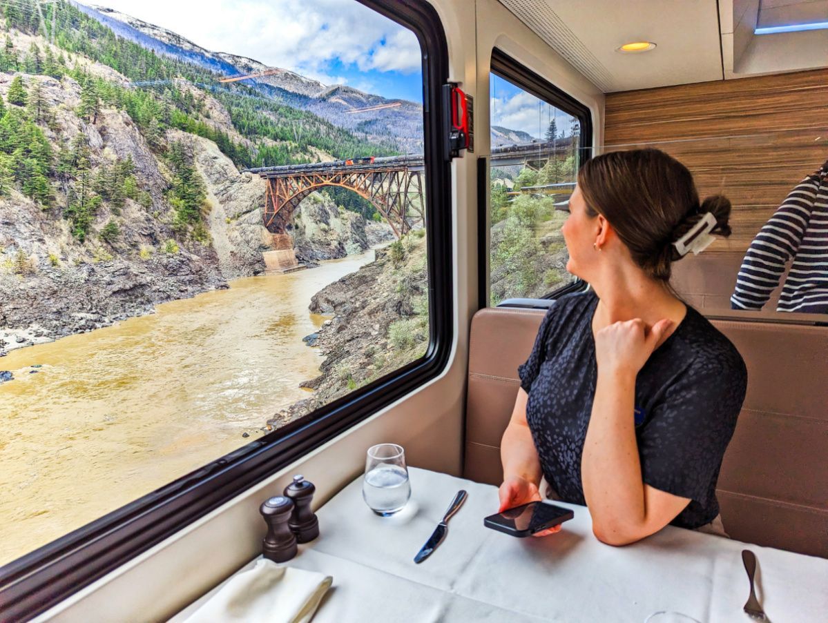 Woman gazing out train window at river, bridge, and mountains. Table with glass, phone, and salt/pepper shakers.