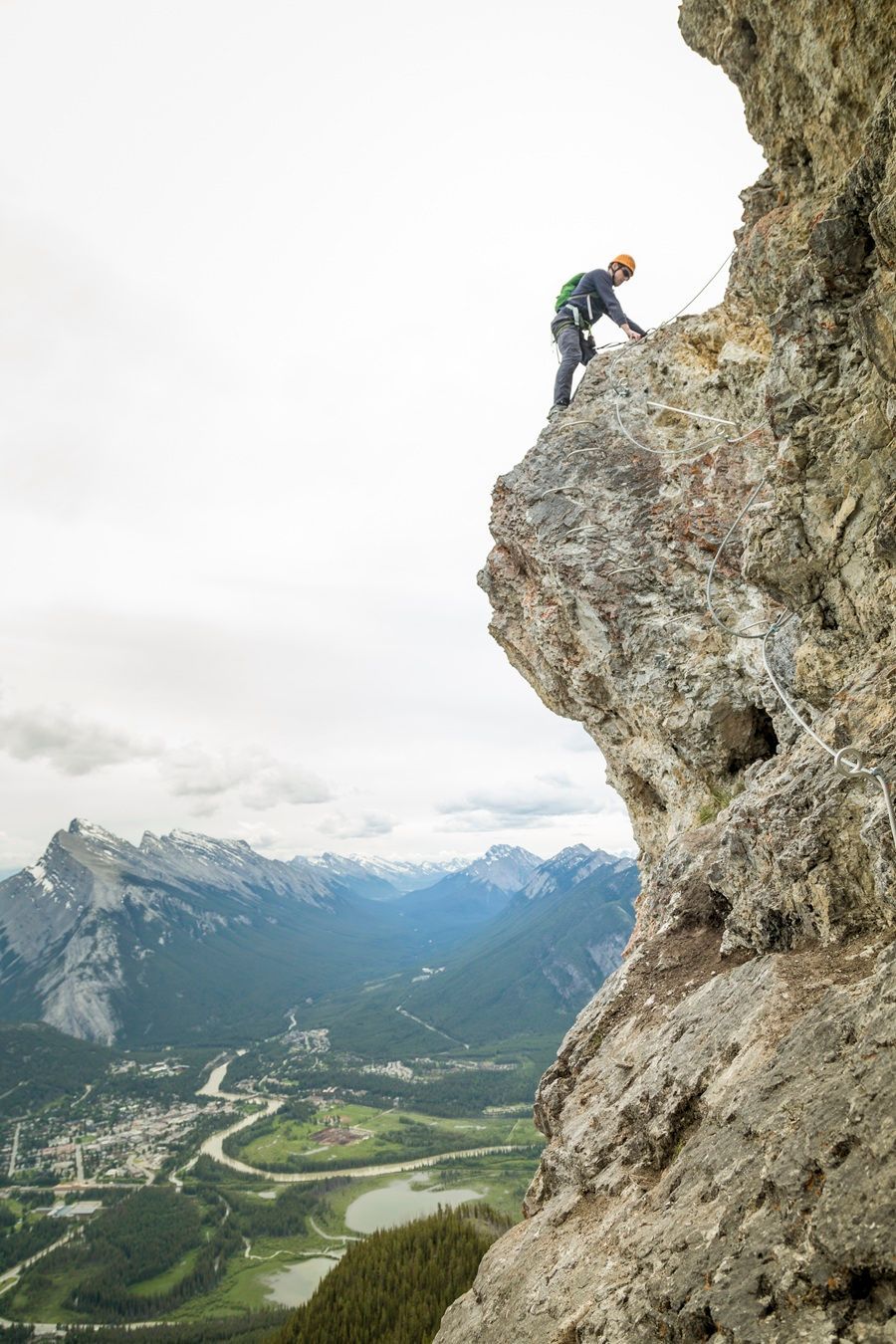 Rock climber scaling a cliff face with a mountainous backdrop and town below.