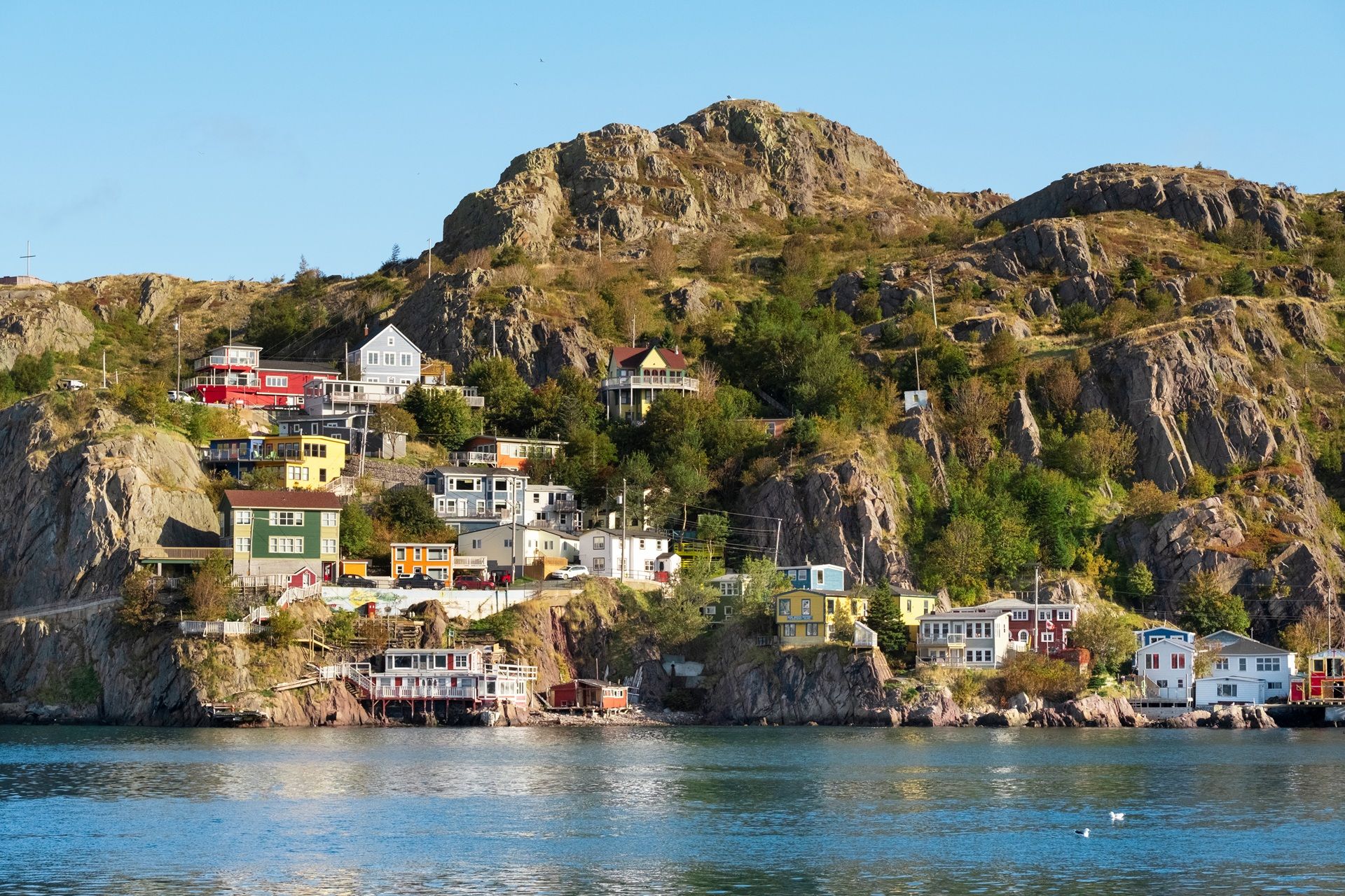 @ Destination Canada Colorful houses built on a rocky hillside overlooking a body of water under a blue sky.