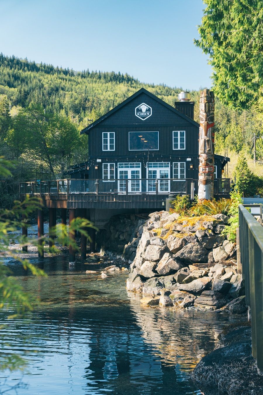 Zwart houten gebouw boven het water, klein balkon, berglandschap op de achtergrond, totempaal, bomen.