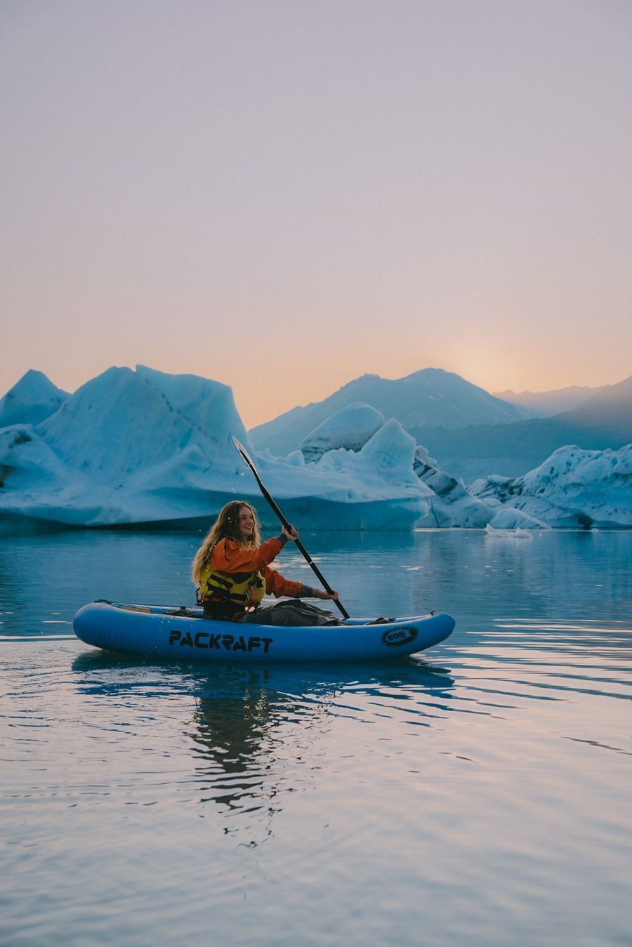 Woman kayaking among icebergs in a blue lake, holding a paddle; sunset in the background.