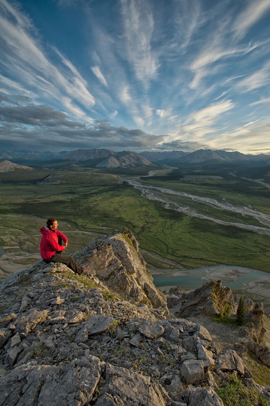 Person in red jacket sits on rocky peak, looking at a valley and sky with streaky clouds.
