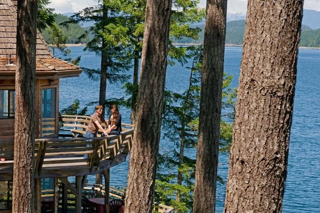 Mensen ontspannen op een terras aan het meer, tussen hoge bomen, met uitzicht op het blauwe water.