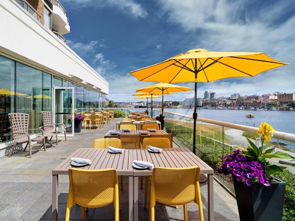 Terras van een restaurant aan het water met gele parasols, rieten stoelen, tafels en uitzicht op de haven.
