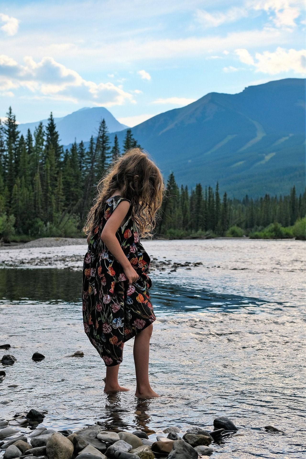 Girl in floral dress wading in a river, mountains and trees in the background.