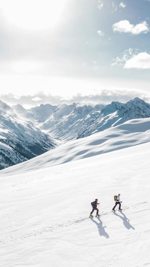Two hikers ascending a snow-covered mountain, sunny day. Mountain range in background.