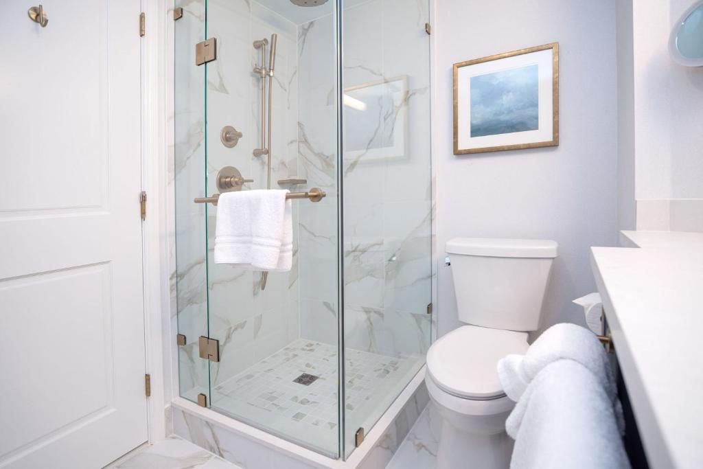 Lichte, witte badkamer met glazen douchecabine, toilet, handdoek en ingelijste muurdecoratie.