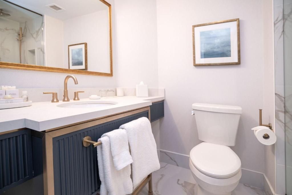 Lichte badkamer met wastafelmeubel, gouden kraan, spiegel, ingelijste kunst, toilet en handdoekenrek.