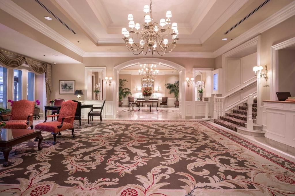Elegante hotellobby met kroonluchter, tapijt met patroon, trappen en rode fauteuils.