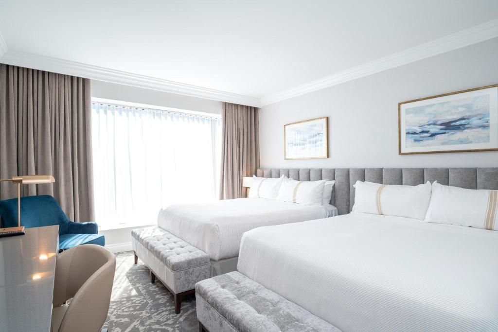 Lichte hotelkamer met twee witte bedden, een blauwe stoel en een groot raam met gordijnen.