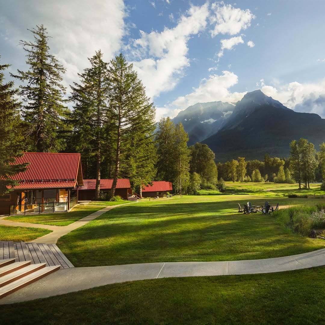 Hutten van Tweedsmuir Park Lodge naast een groen gazon en hoge dennenbomen, met een bewolkte bergtop op de achtergrond.