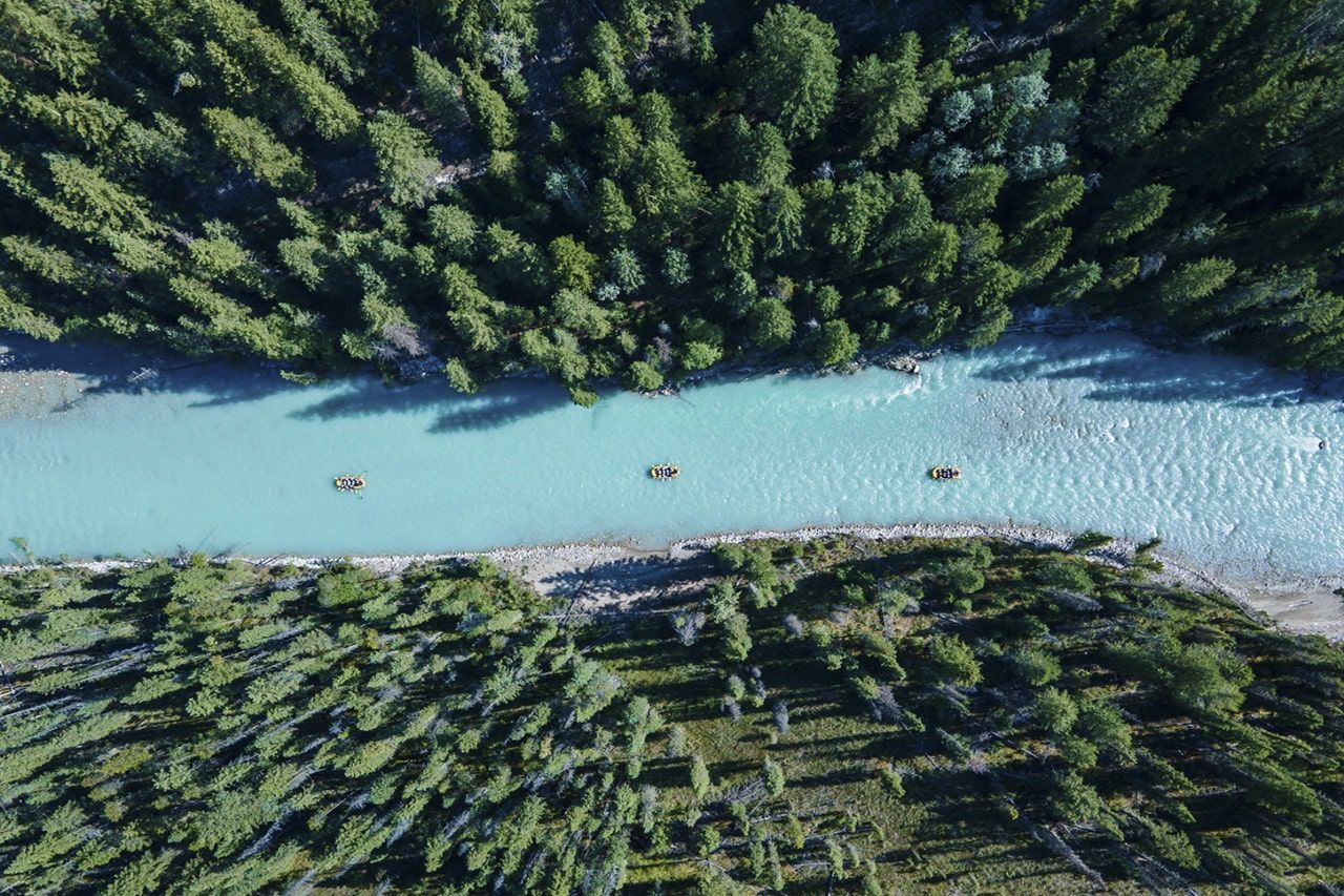 Luchtfoto van een turquoise rivier die door een dicht groen bos stroomt, met drie kleine bootjes op het water.