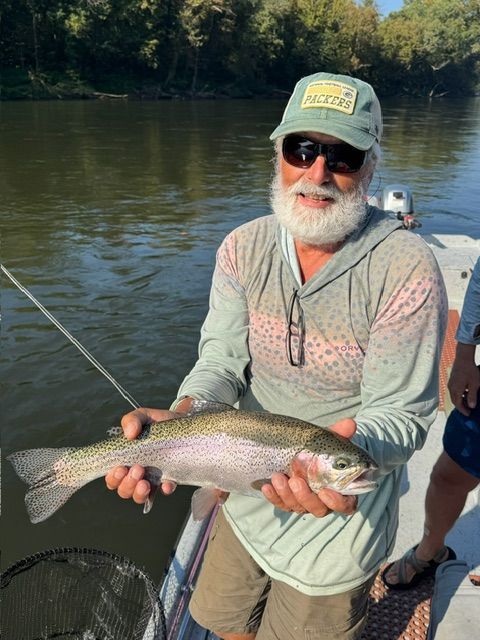 Man in hat and sunglasses holding a rainbow trout on a boat, green river in background.
