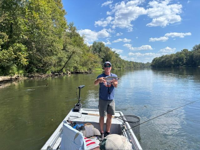 Man on a boat holds up a caught fish on a river. Sunny day, trees on banks, blue sky.