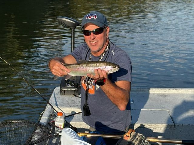 Man in sunglasses holding a rainbow trout, smiling in a boat on a calm river.