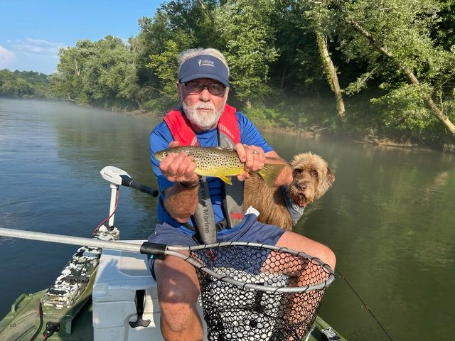 Man in blue shirt, life vest, holding a fish; dog nearby on a boat; misty river.