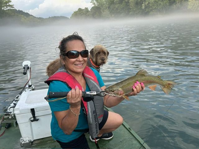 Woman in life vest, holding a large fish on a boat, with dog behind her. Misty river background.