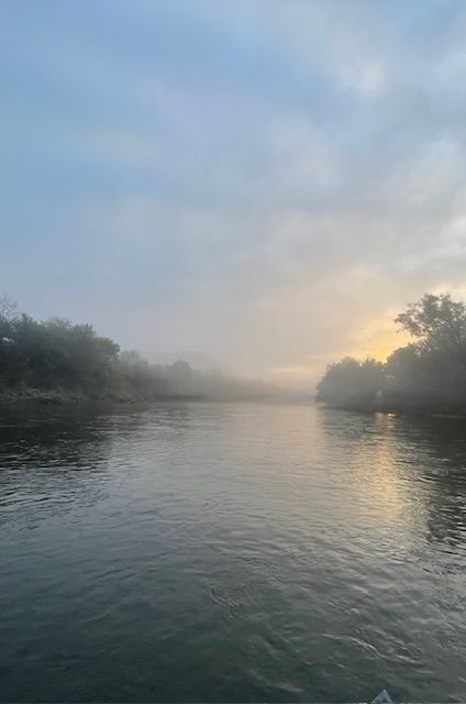River at dawn, shrouded in mist, trees lining the banks, soft golden light breaking through the fog.
