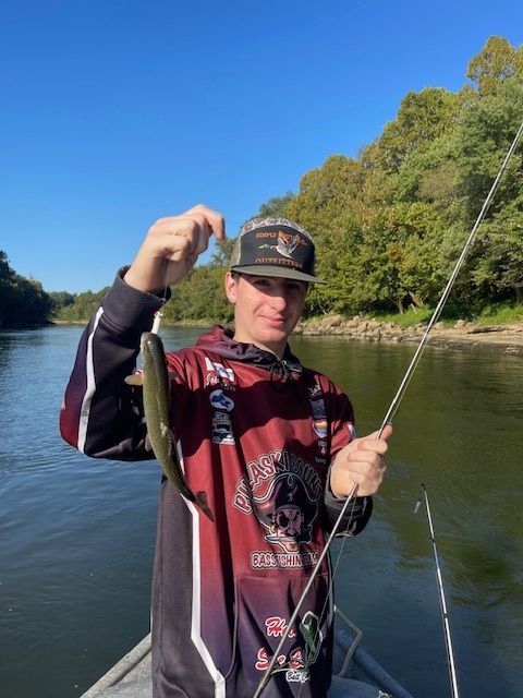Person holding up small fish with fishing rod on a boat in a river.