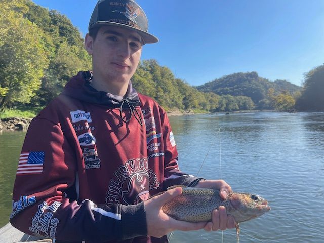 Young person holding a fish, standing near a river with hills in the background on a sunny day.