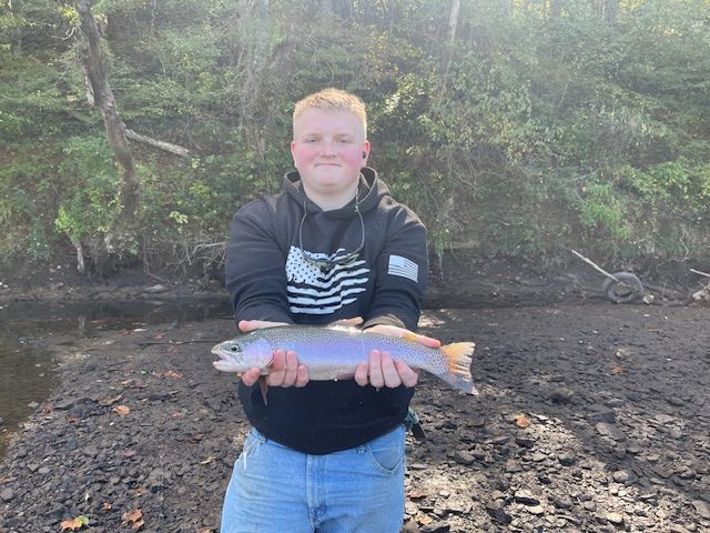 Man holding a fish by a river. He's wearing a black hoodie and jeans. Brown water and trees in the background.