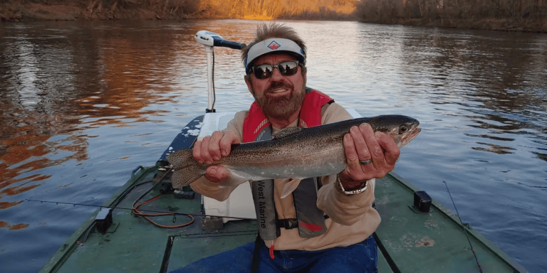 Man in a boat holds up a large fish. Brown water, trees in the background. Man smiles and wears a visor and life jacket.