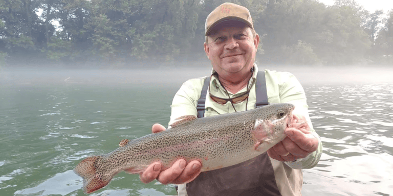 Man in a cap, holding a large rainbow trout in a misty river.