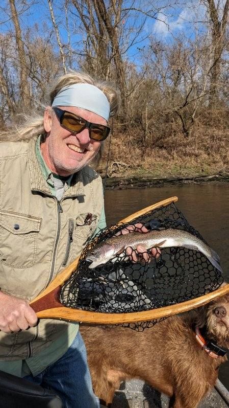 Man holding a fish in a net on a boat, smiling. Brown dog. Outdoors, sunny day.