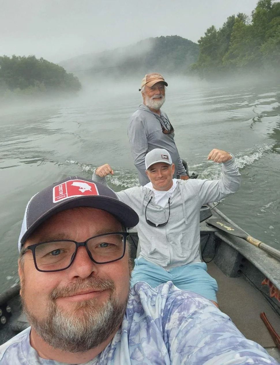 Three men on a boat in a misty river, one flexing, another smiling, one taking a selfie.