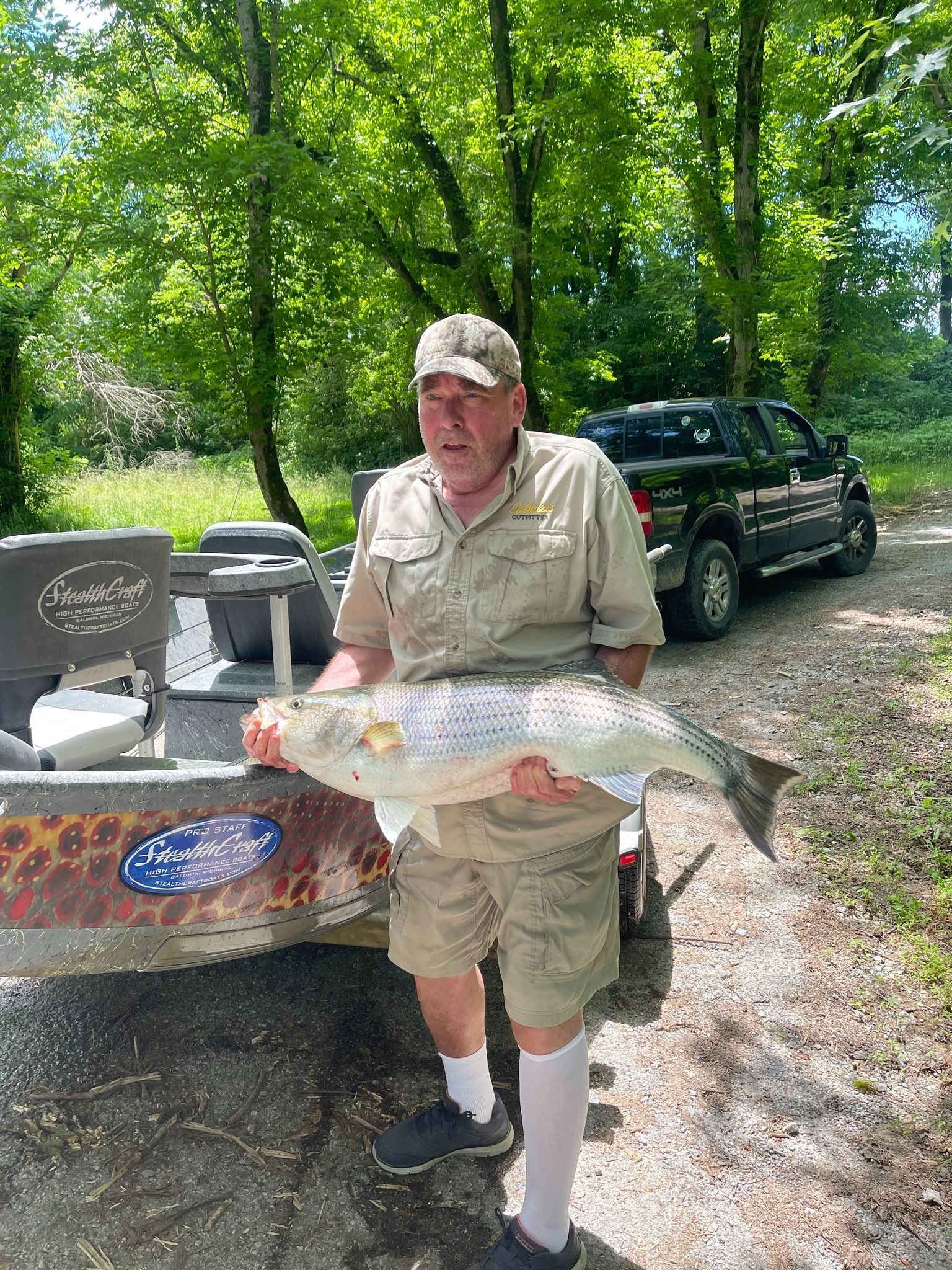 Man holding large fish next to a boat and truck outdoors.