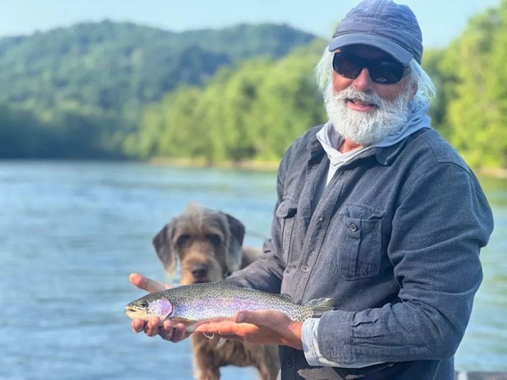 Man holding a rainbow trout, smiling by a lake with a dog.