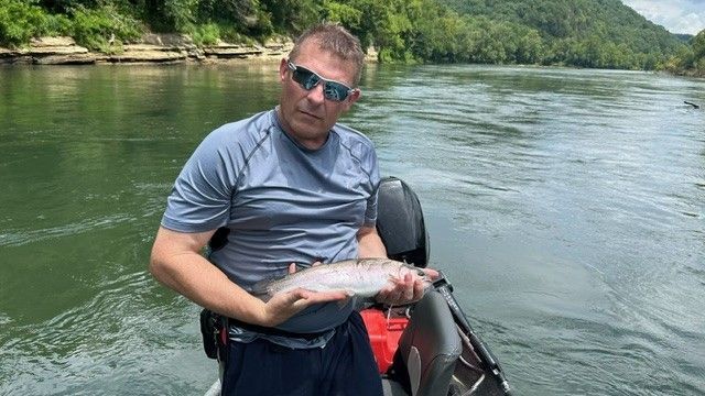 Man holding a fish on a boat in a river. He wears sunglasses and a gray shirt. Green trees in the background.