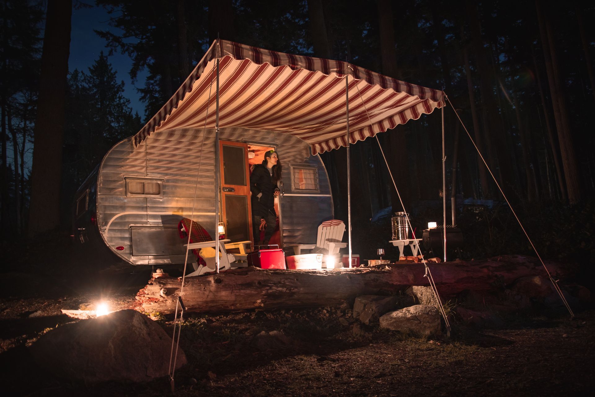 Lit campsite with a striped canopy tent, chair, and supplies beside a camper in a dark forest.