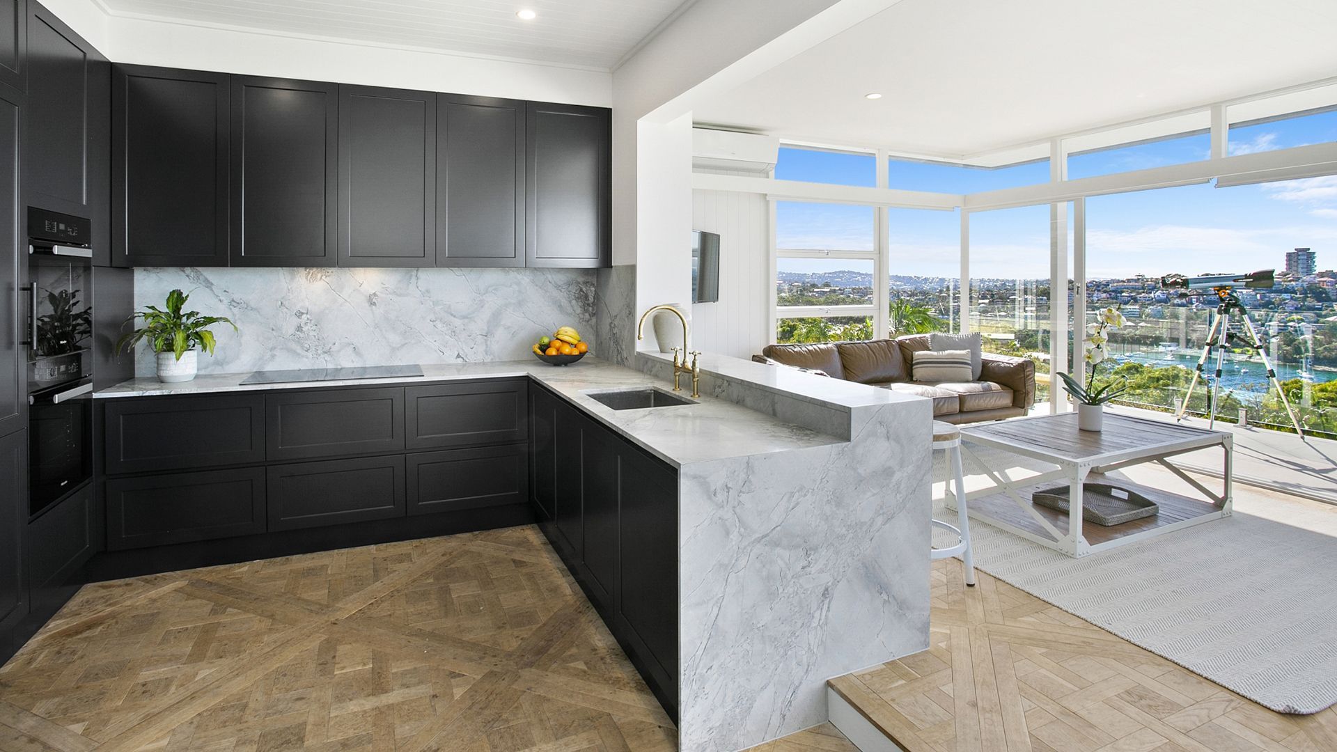 A kitchen with black cabinets and marble counter tops is open to a living room.