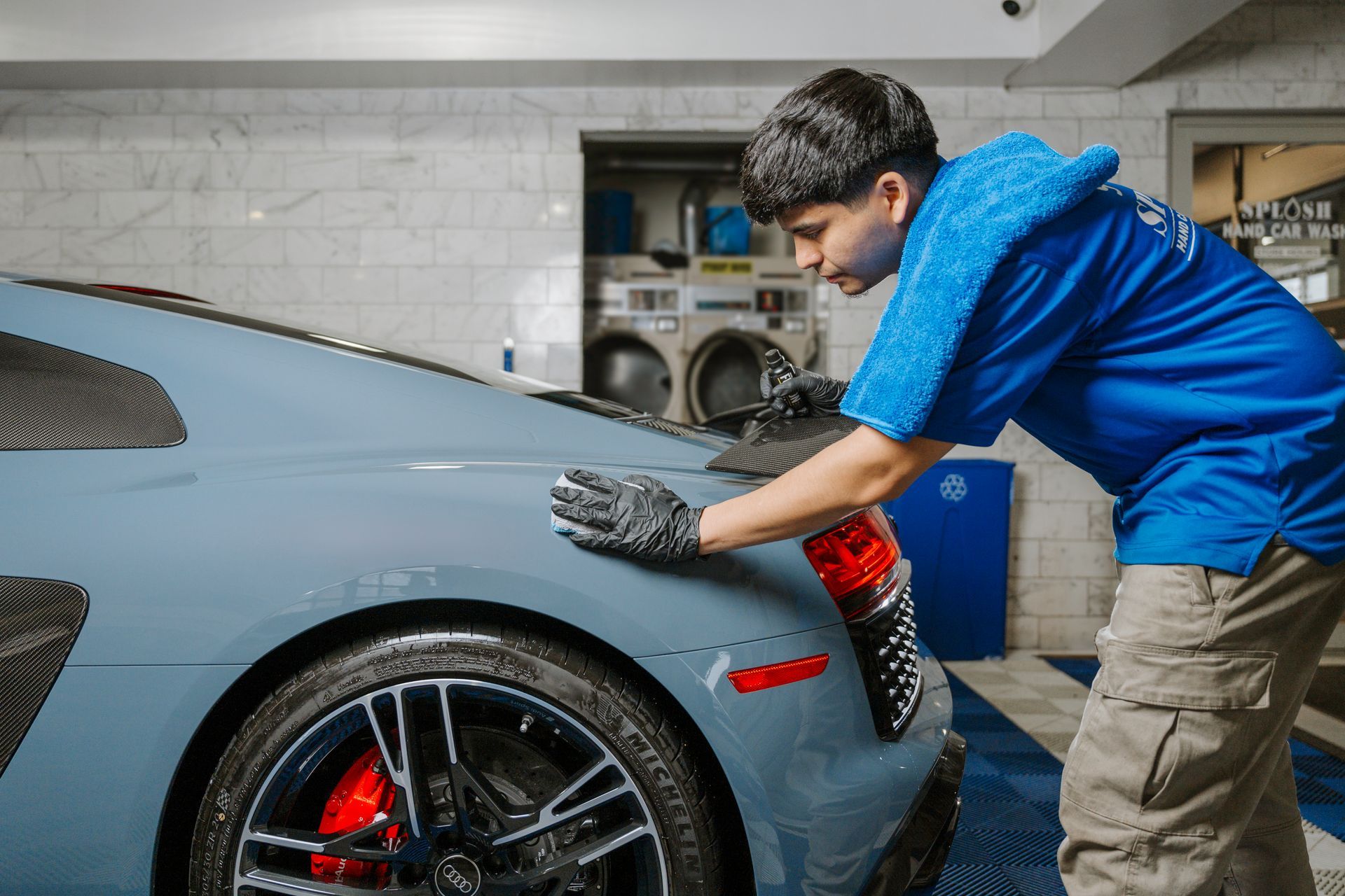 Man in blue shirt and gloves polishing a blue sports car in a garage.