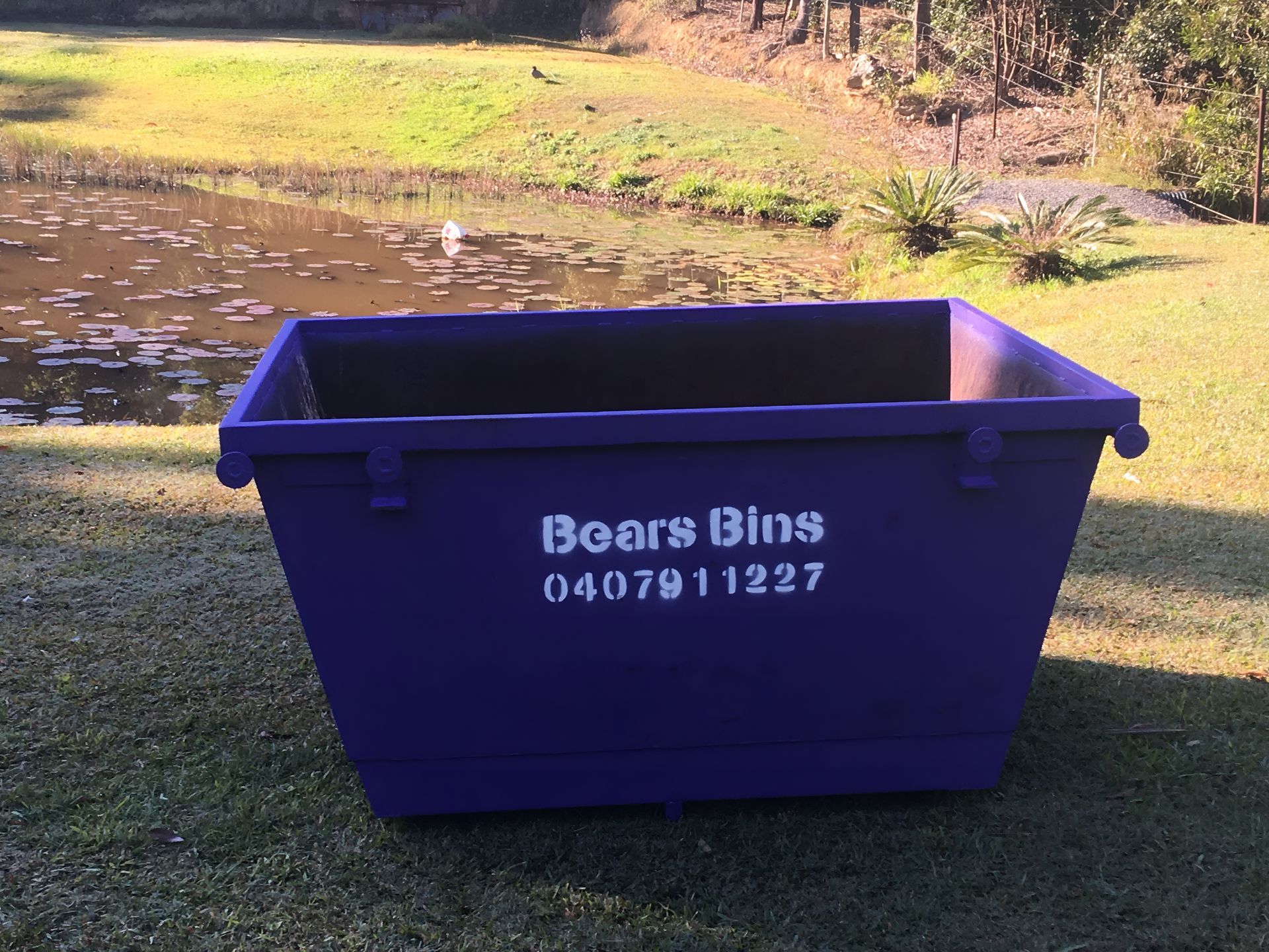 Purple Boars Bins Dumpster on Grass, Near Pond — Skip Bin Hire Sunshine Coast in Cooroy, QLD