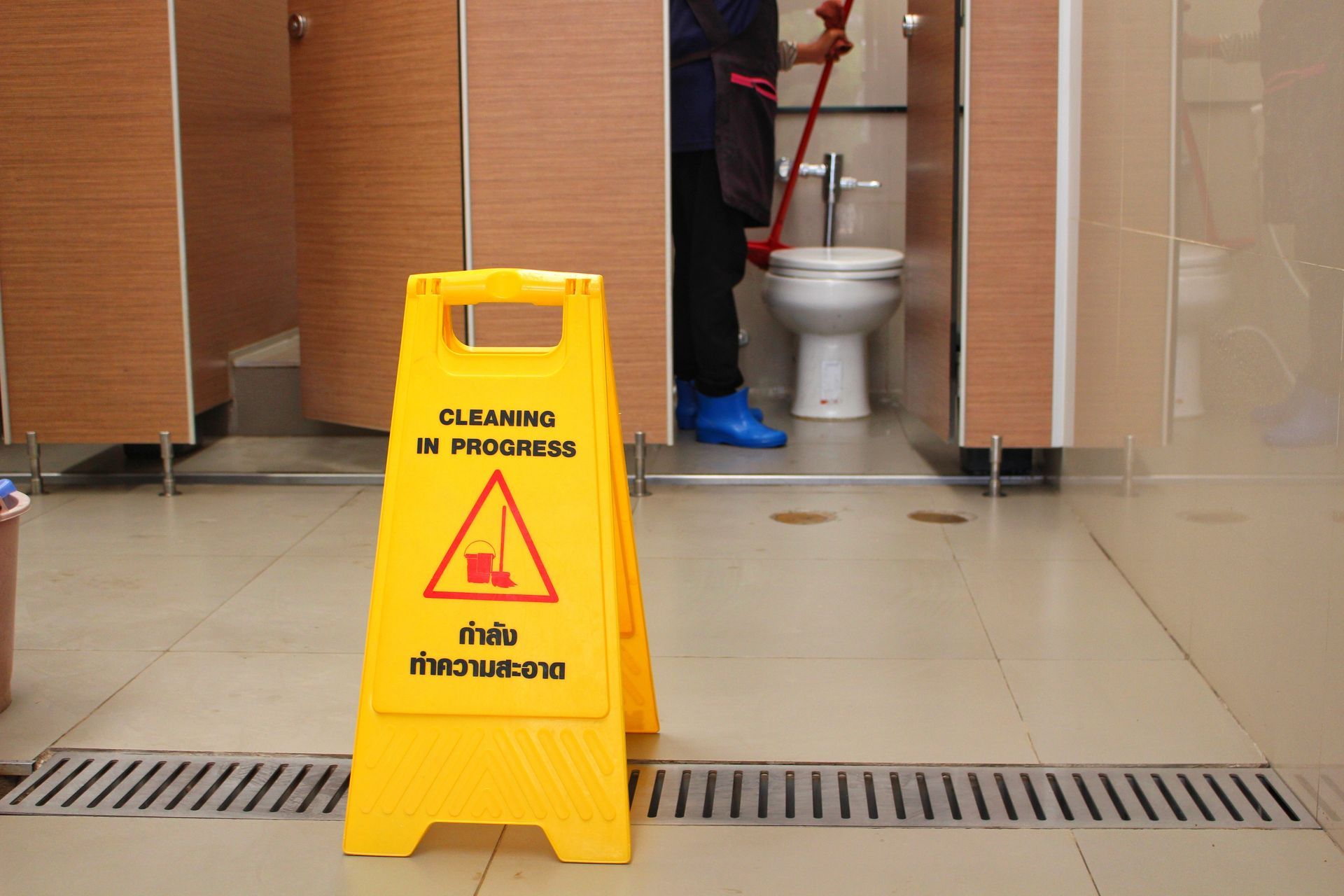 Someone cleaning the toilets in the bathroom of an office building. A cleaning in profess sign is on display to warn guests.