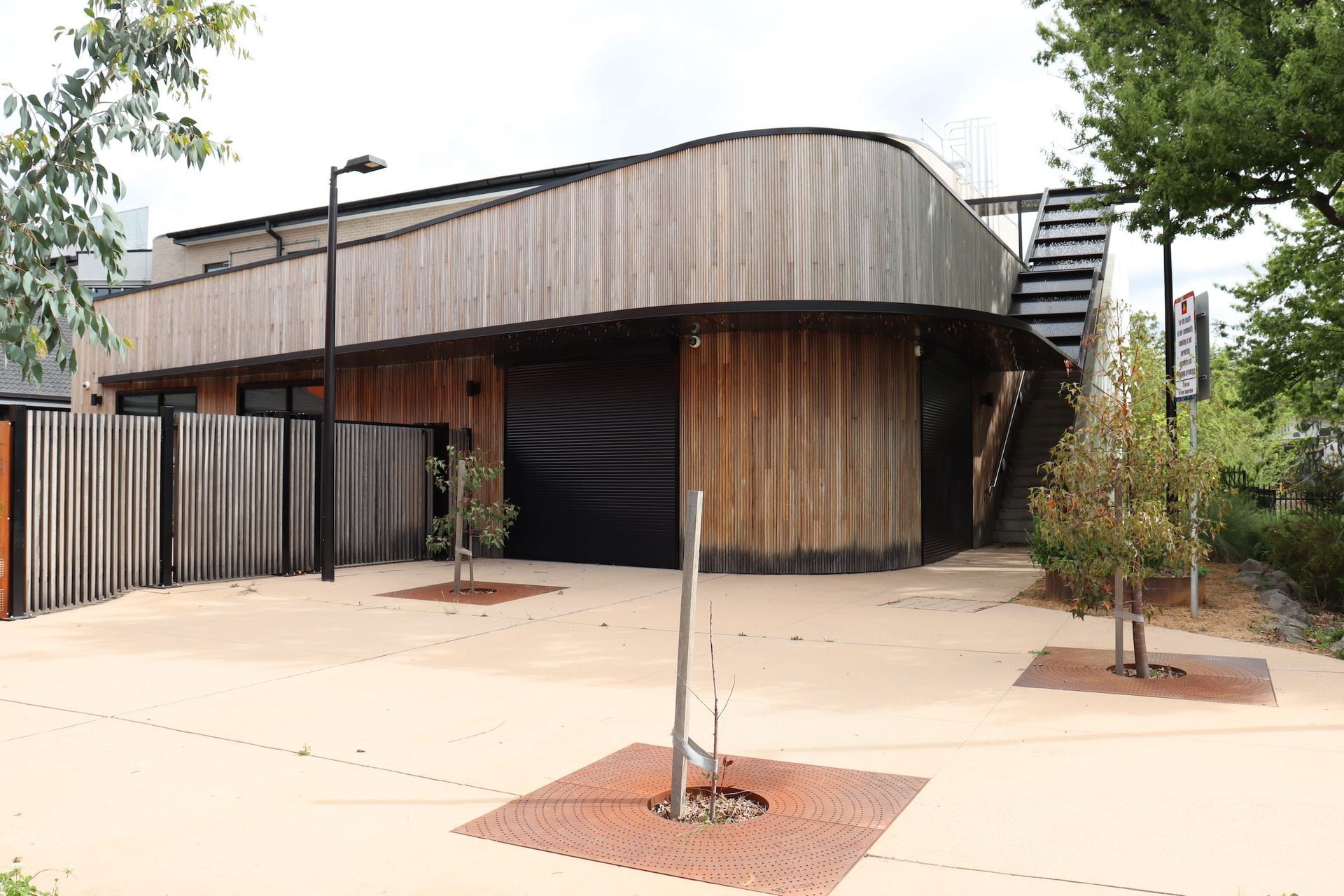 A large wooden building with a curved roof is surrounded by trees and a fence.