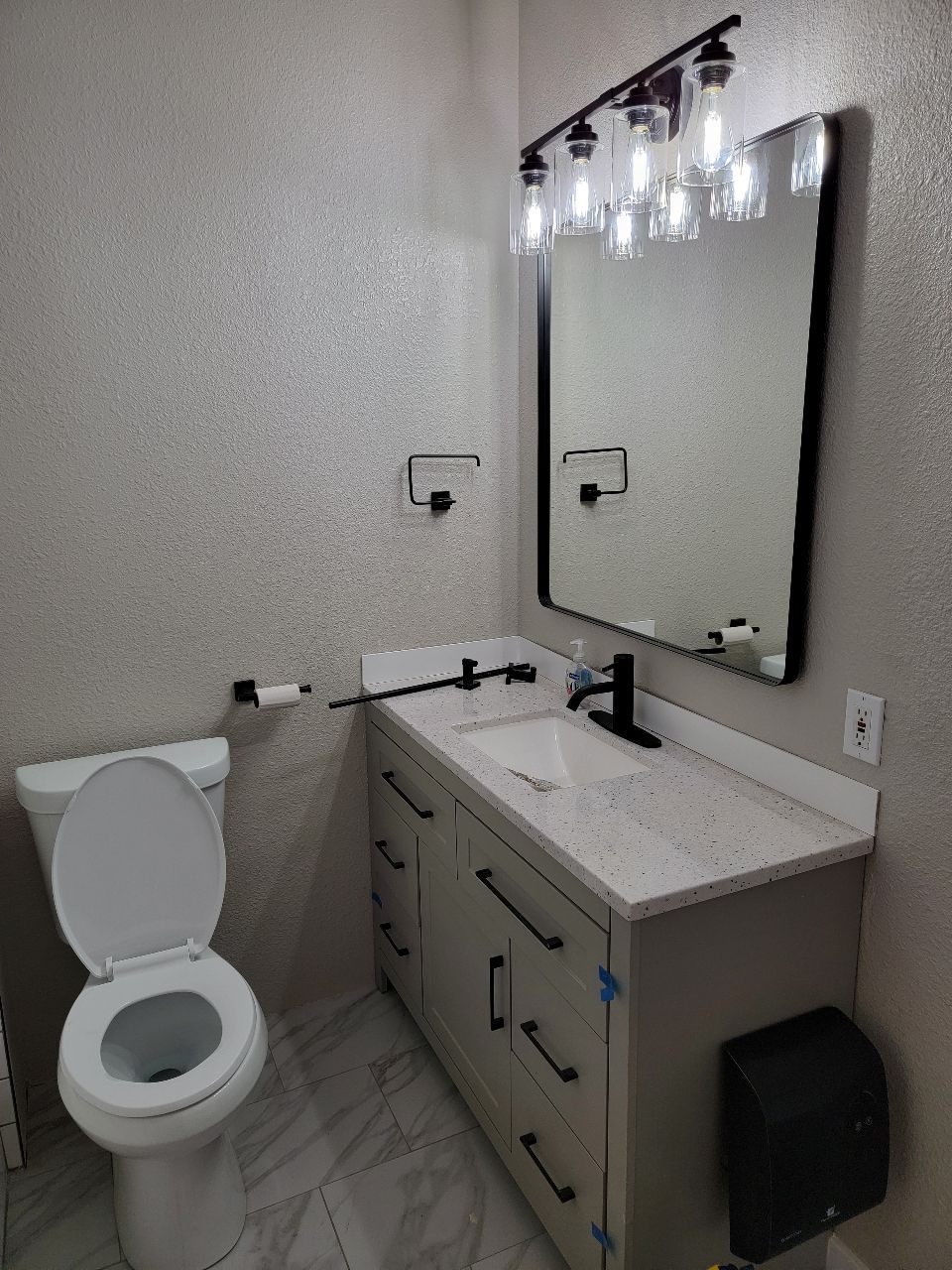 Bathroom with toilet, vanity, mirror, and black fixtures. Light-colored walls and floor.