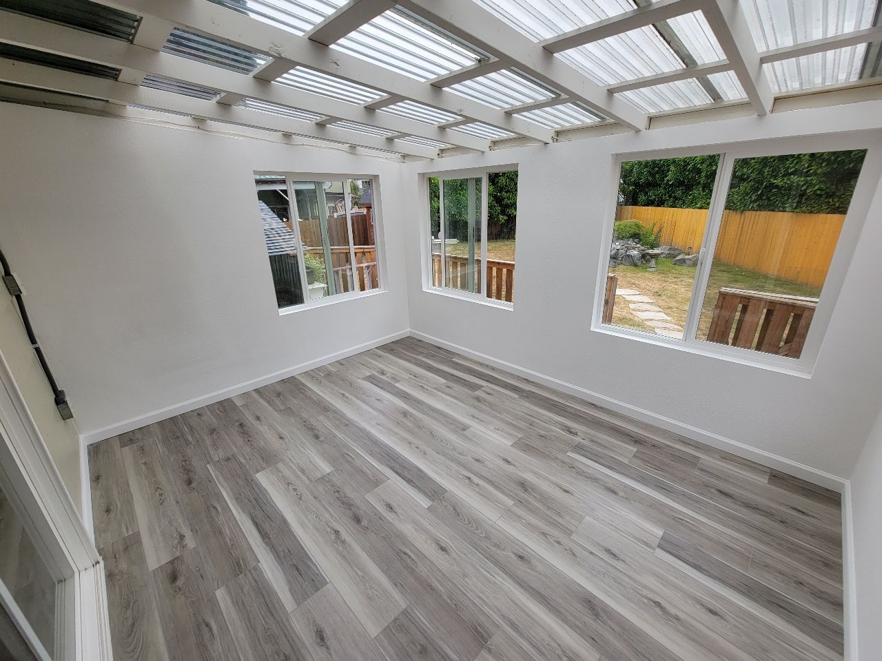 Interior view of a small room with white walls, gray wood-look floor, and windows; partially translucent roof.